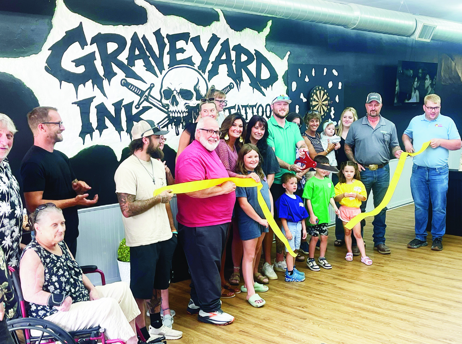 Cooper Blackledge, center with scissors, along with friends and family celebrated the opening of Graveyard Ink tattoo shop with a ribbon cutting on Friday. The group is standing in front of a graphic that was created by Korina Martin.